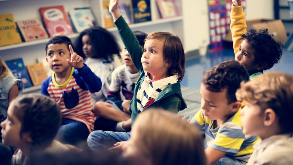 A group of children in casual clothes sitting on a carpet and looking forwards, some children have their hand up to answer a question