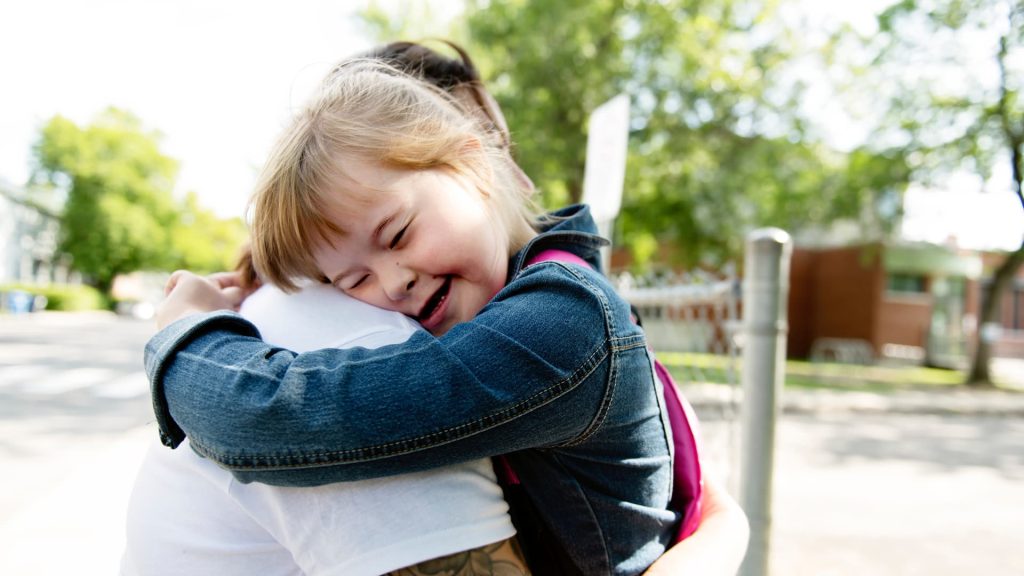 Young girl wearing denim shirt hugs adult on a school playground.