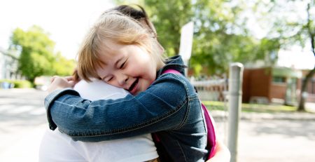 Young girl wearing denim shirt hugs adult on a school playground.
